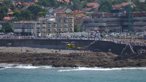 gijon, spain - july 27, 2025: a yellow fire helicopter lowers a water bucket near the rocky coastline of gijon during firefighting operations. the aircraft operates close to the beach while crowds observe