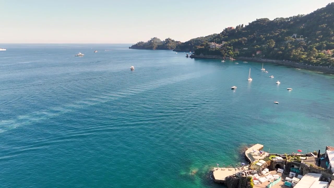 Ripresa aerea con drone cattura acqua turchese limpida e lussuosi edifici costieri sotto la luce solare intensa.