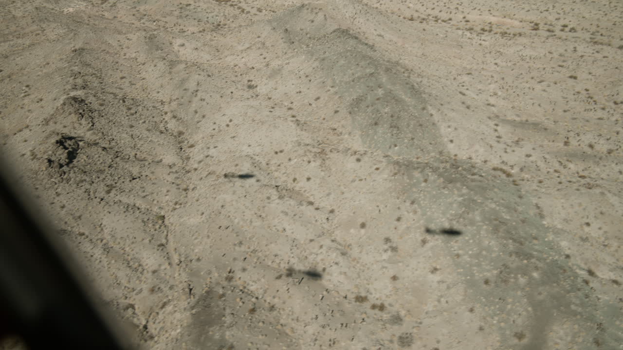 Helicopters fly above a vast, arid landscape showcasing intricate landforms and shadowy contours. The rugged terrain features dry hills and scattered vegetation, highlighting nature's beauty.
