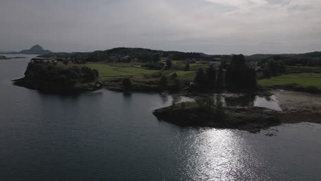 sunlight reflection on calm waters of klubboya island in bronnoysund, norway with green field at the coast. aerial orbit