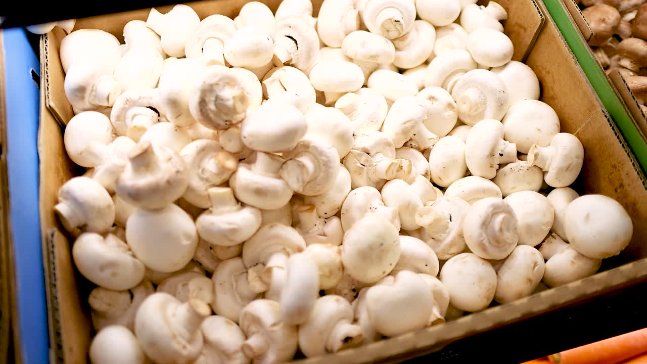 A hand selects white button mushrooms from a supermarket display under bright lighting. The setting is Gold Coast, Australia