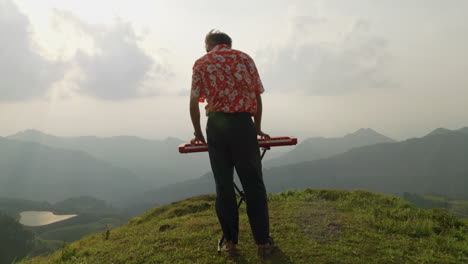 man faces away from the camera on a hilltop and plays the piano to the valley floor below