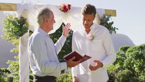 Lächelnder-Kaukasischer-älterer-Männlicher-Hochzeitsoffizier-Mit-Buch-Und-Bräutigam,-Der-Im-Außenaltar-Steht