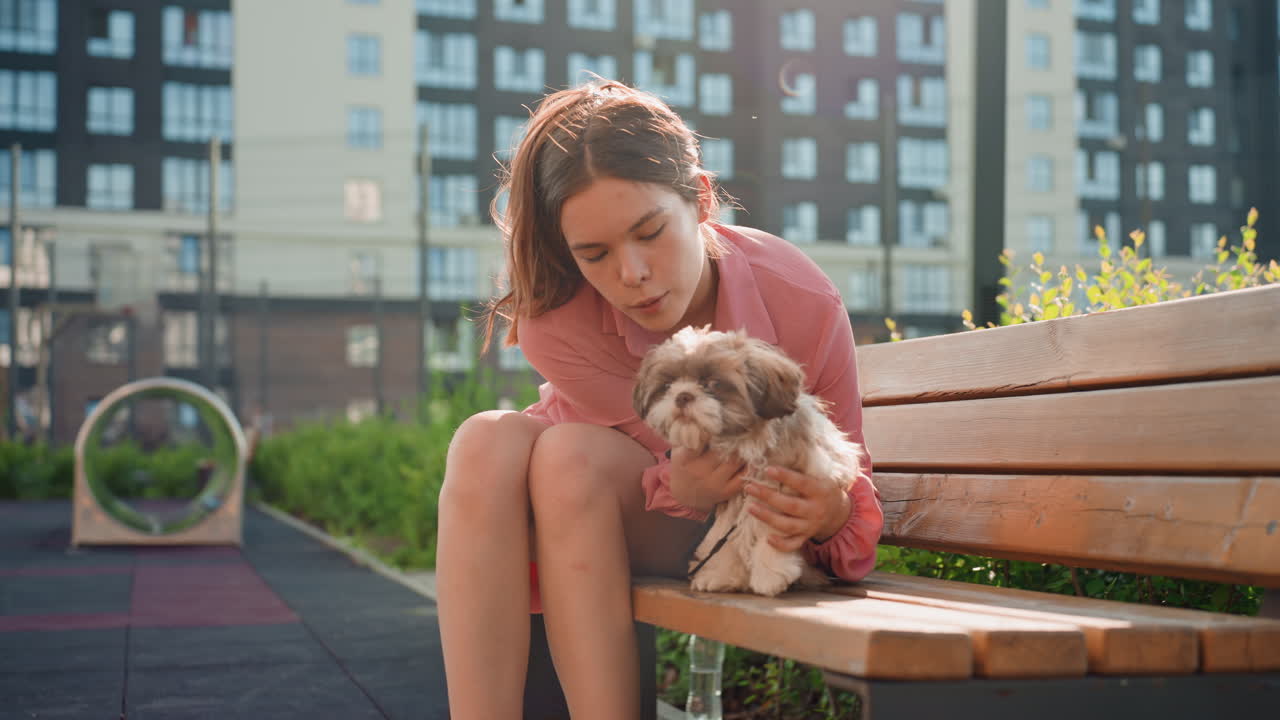Caucasian Woman Enjoying Puppy In Park, Woman Seated On Bench Affectionately Playing With Puppy Outside, Bright Sunny Afternoon Captures Woman Joyfully Cuddling Her Puppy Near Urban Apartments