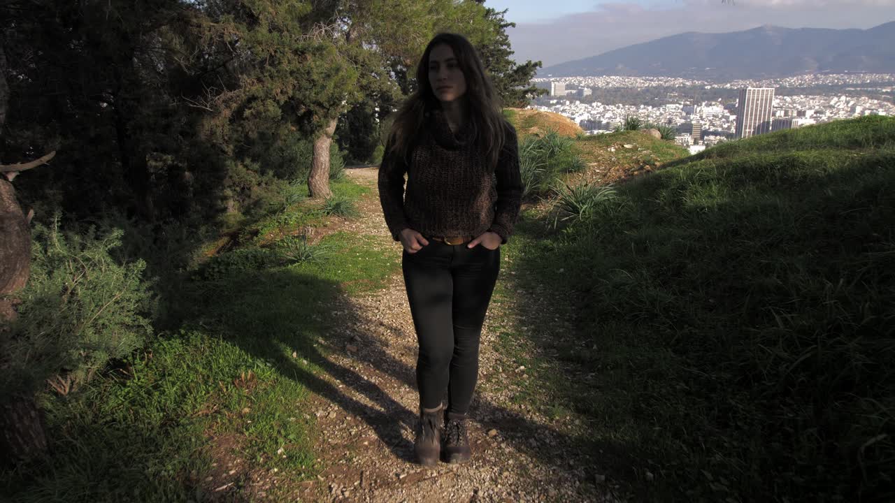 Slow motion gimbal, young woman walking on path having hands in pockets on Lycabettus hill, city of Athens in the background