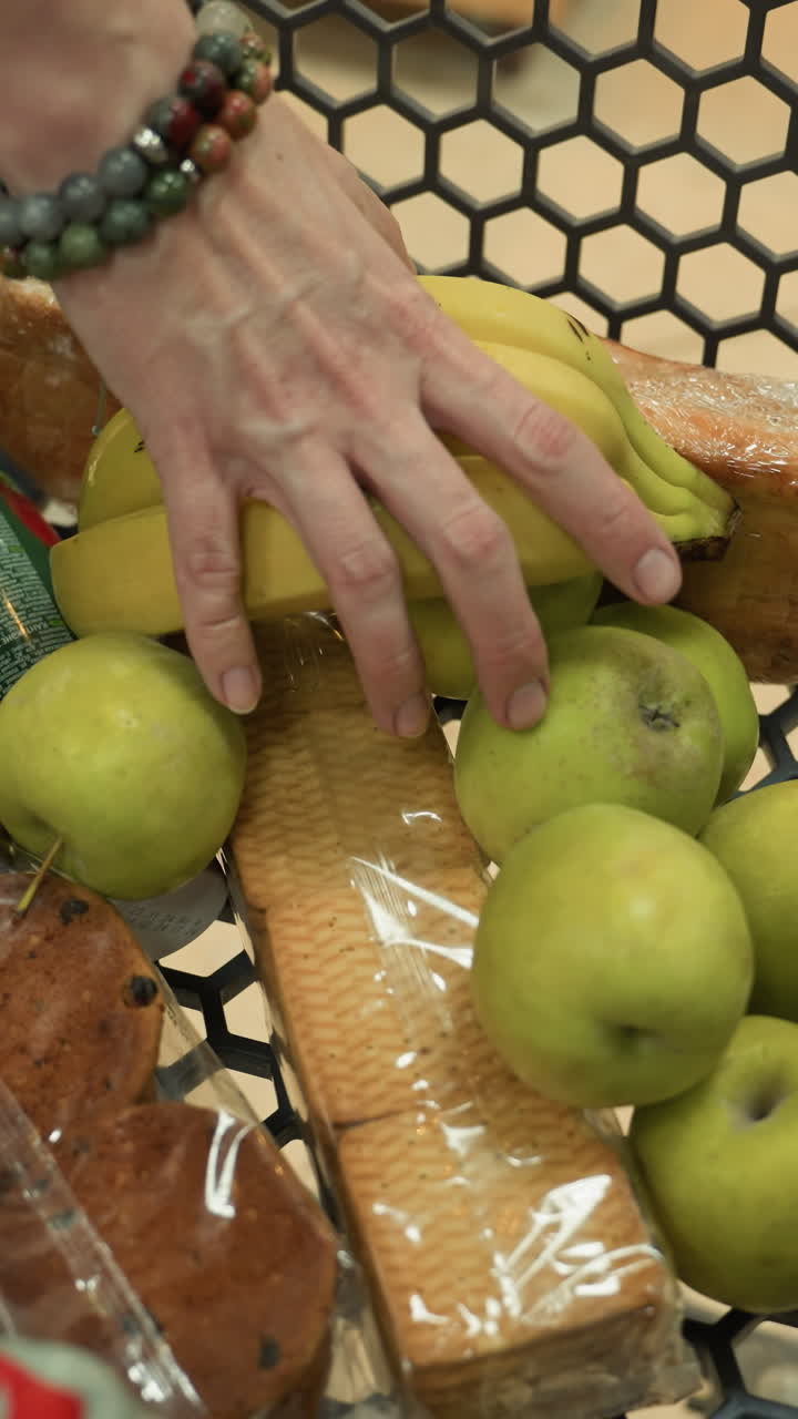 una dama con un abrigo negro y botas negras colocando plátanos en un carrito de compras que contiene un paquete de galletas, un pequeño jugo, una panada de pan y siete manzanas
