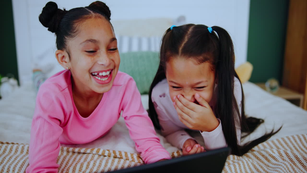 Two young girls laughing together while using a tablet in bed