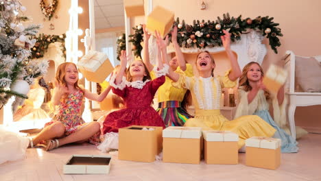 adorable teenage girls in cute dresses toss the gift boxes happily. kids sit at the festively decorated fireplace in photo studio. christmas decor.