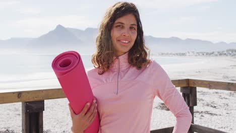 portrait of healthy mixed race woman exercising outdoors holding yoga mat standing by the sea