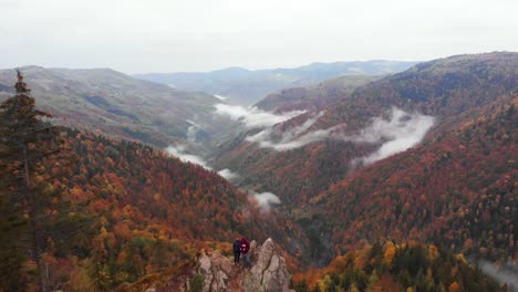 Pareja-De-Vista-Aérea-Se-Paran-En-La-Cima-De-Una-Roca-Cerca-Del-Valle-De-La-Montaña,-Otoño