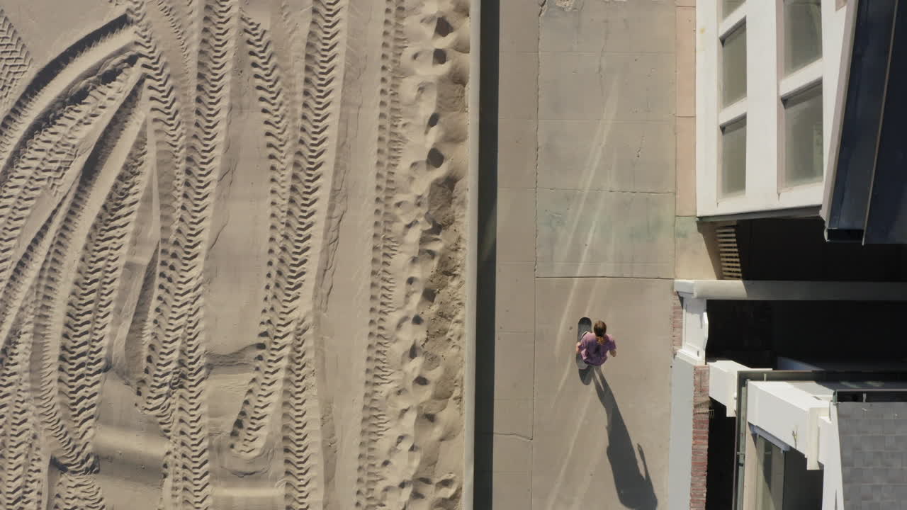persona montando monopatín en un camino de hormigón cerca de la playa de arena, vista aérea de arriba hacia abajo