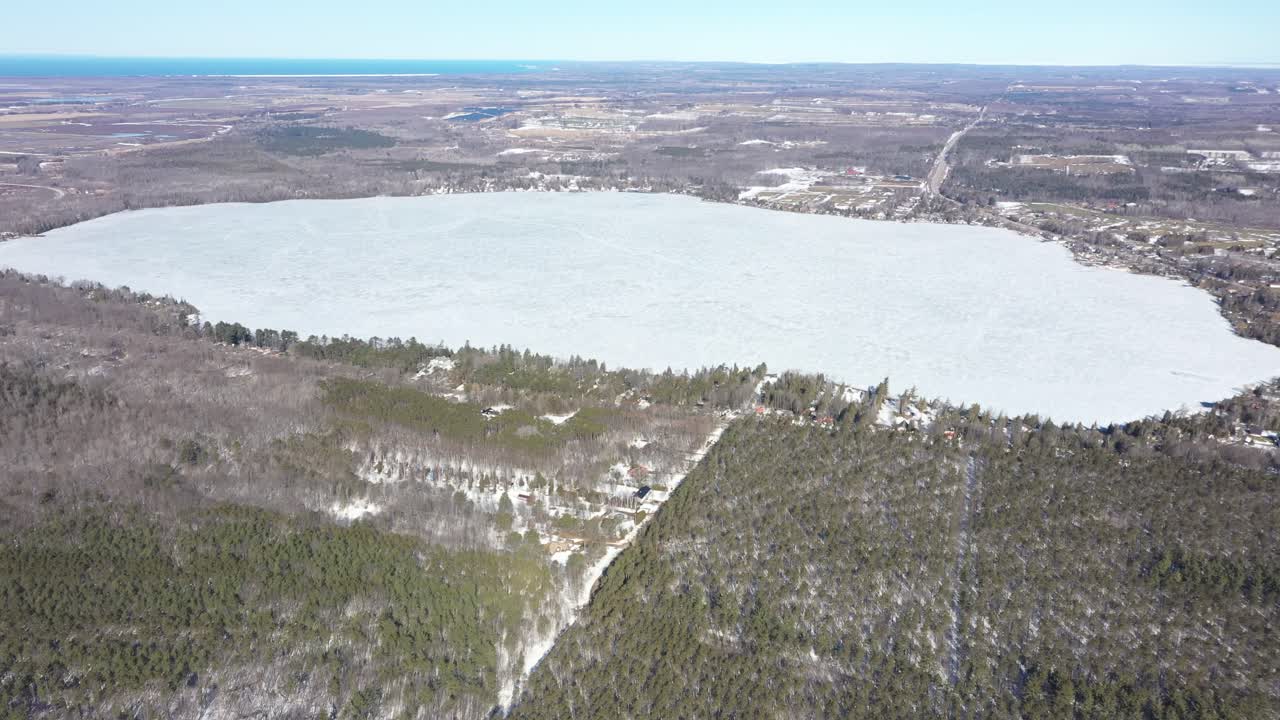 Ilmakuva, jossa liikutaan eteenpäin ja kallistetaan ylöspäin laajan jäätyneen järven ja lumipeitteisen ikivihreän metsän yllä kirkkaana, aurinkoisena talvipäivänä, paljastaen kaukaisen horisontin ja maiseman