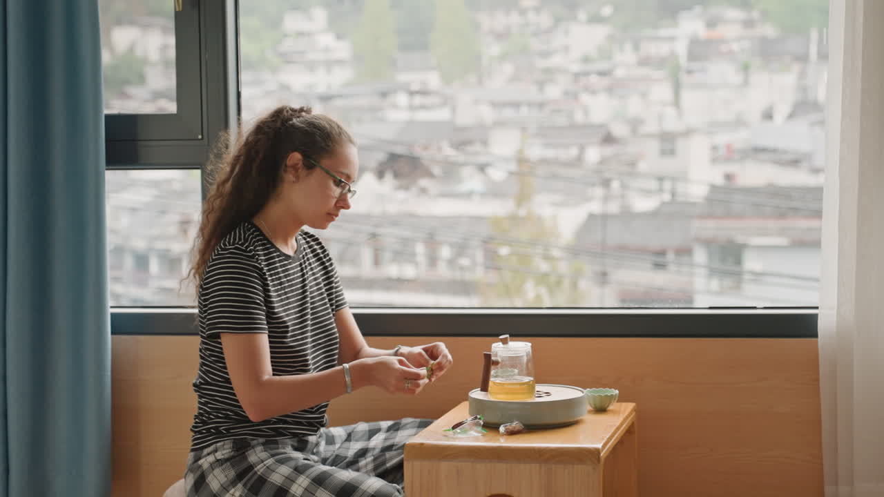 Hotel breakfast ritual woman preparing tea and snack on compact bedside table, careful hand movements spreading jam on toast, porcelain cup and teapot, calm city backdrop through window, intimate
