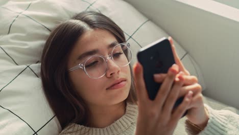 Close-up-of-caucasian-teenage-girl-browsing-phone-while-lying-on-bed-on-her-back