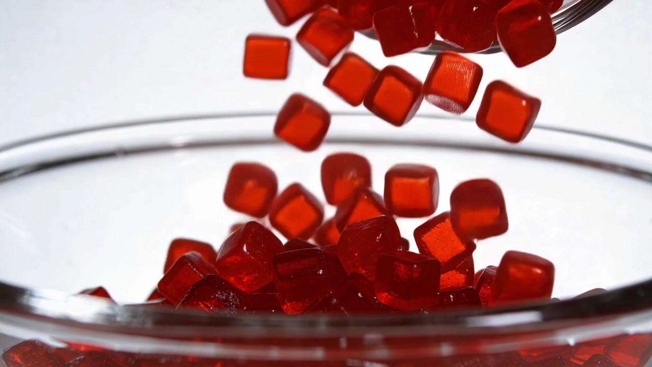 Pouring metal spoon releasing red gel cubes into clear glass bowl in studio, white backdrop