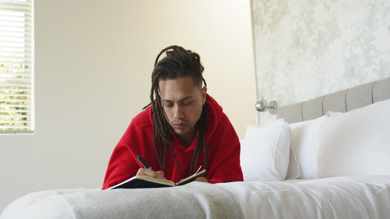 Mann mit rotem Hoodie sitzt, hält inne, um seine Gedanken zu sammeln, und schreibt in einem Spiralblock im Schlafzimmer