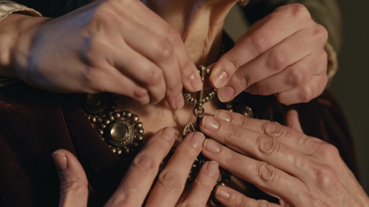 Mãos estendidas fechando o fecho de um colar em uma mulher de vestido bordô, no camarim, para um evento.