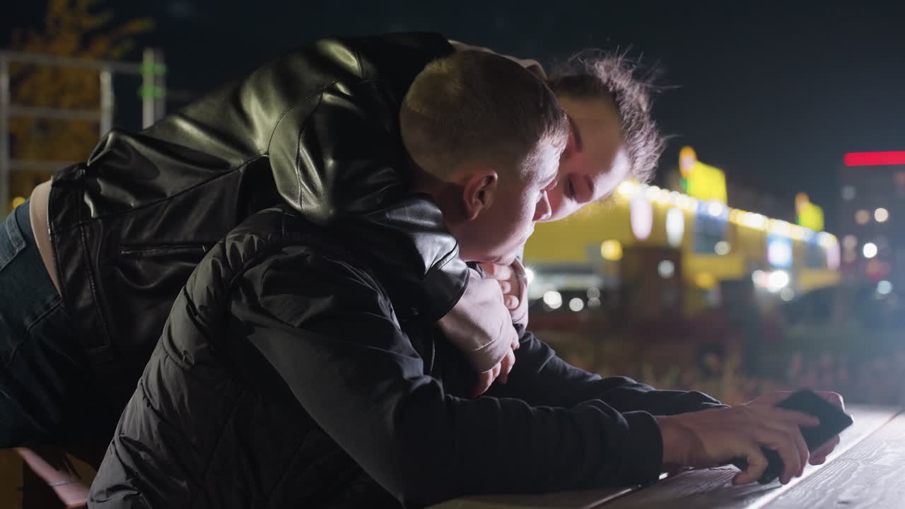 Man focused on phone while seated at wooden table during evening as fiance approaches from behind and gently holds him under vibrant city lighting and urban architecture in nighttime setting