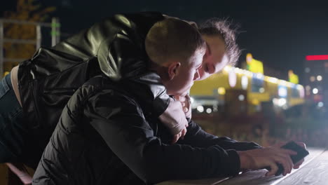 man focused on phone while seated at wooden table during evening as fiance approaches from behind and gently holds him under vibrant city lighting and urban architecture in nighttime setting