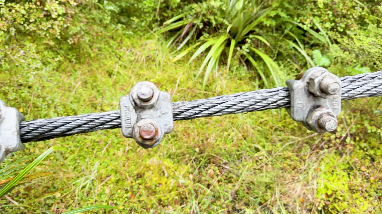 Close-up of a metal cable bridge with bolts and nuts amidst vibrant foliage, showcasing structural details and natural surroundings