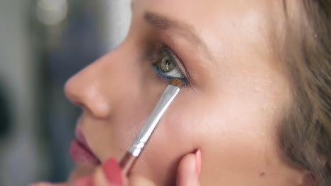 side view of a model's face. make up artist applying eyeshadows under the lower eyelid with brush. blue eyeliner. make up