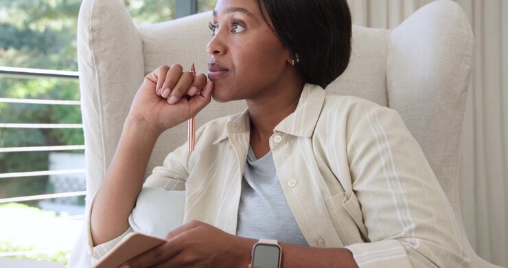 African American woman resting chin then lifting pen by window in armchair, journaling in notebook