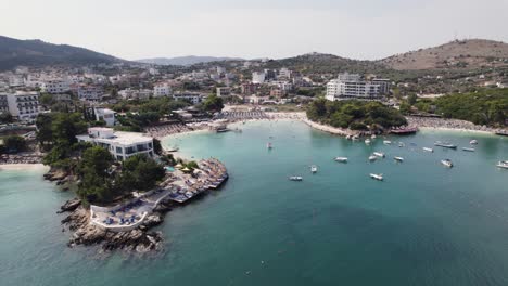 Vista-Aérea-Del-Pintoresco-Pueblo-De-Ksamil-En-La-Costa-Del-Mar-Jónico-De-La-Riviera-Albanesa,-Mostrando-Barcos-En-El-Tranquilo-Puerto