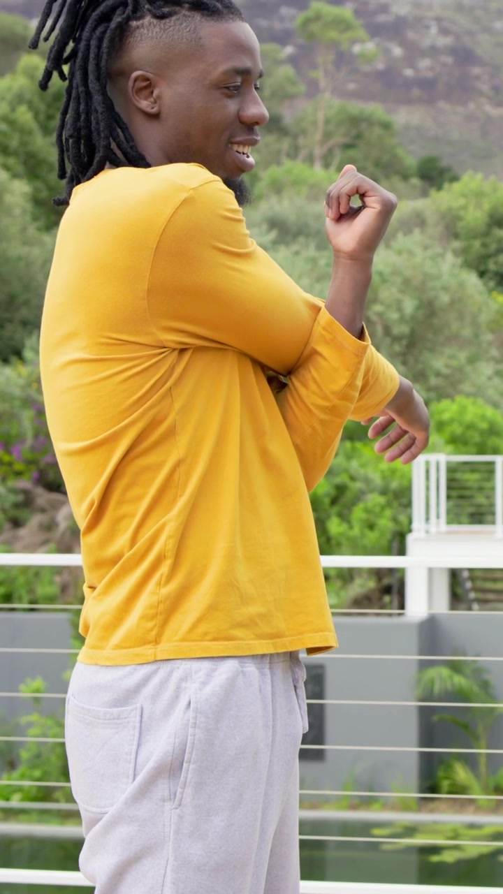 Vertical video: Pausing, man in yellow shirt stretching arms to relax on deck, railing and hillside
