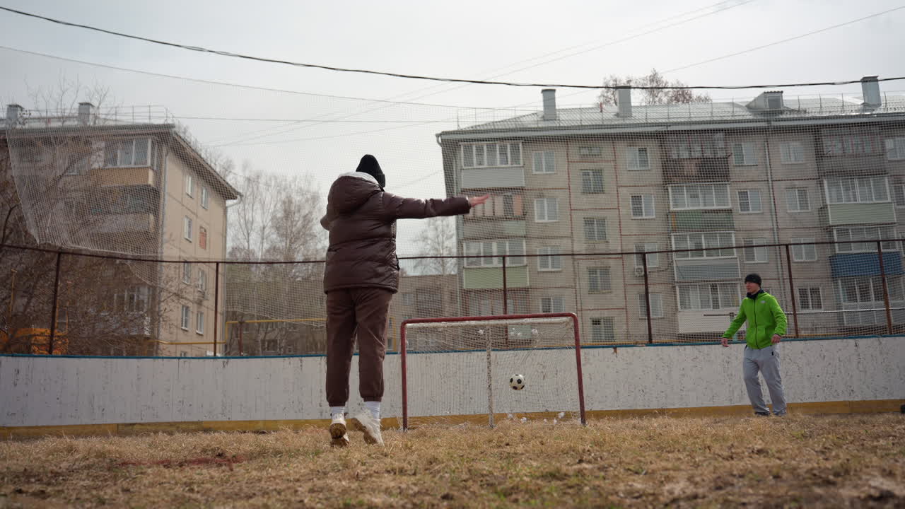 Una mujer blanca patea una pelota de fútbol en un campo urbano, con una chaqueta acolchada marrón y un gorro, apuntando a una portería de metal oxidado mientras el portero, con una chaqueta verde, observa en un día frío y nublado con bloques de apartamentos al fondo.
