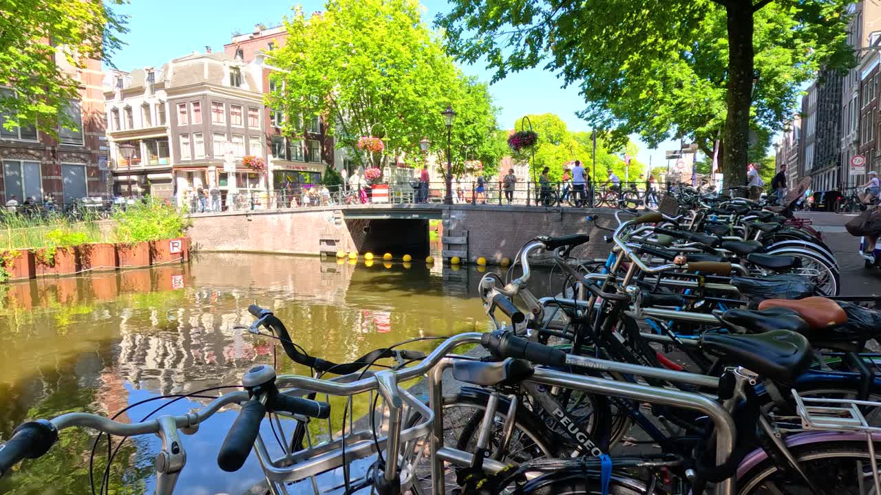 Rows of bicycles beside canal, historic buildings, and trees under bright daylight, wide static shot