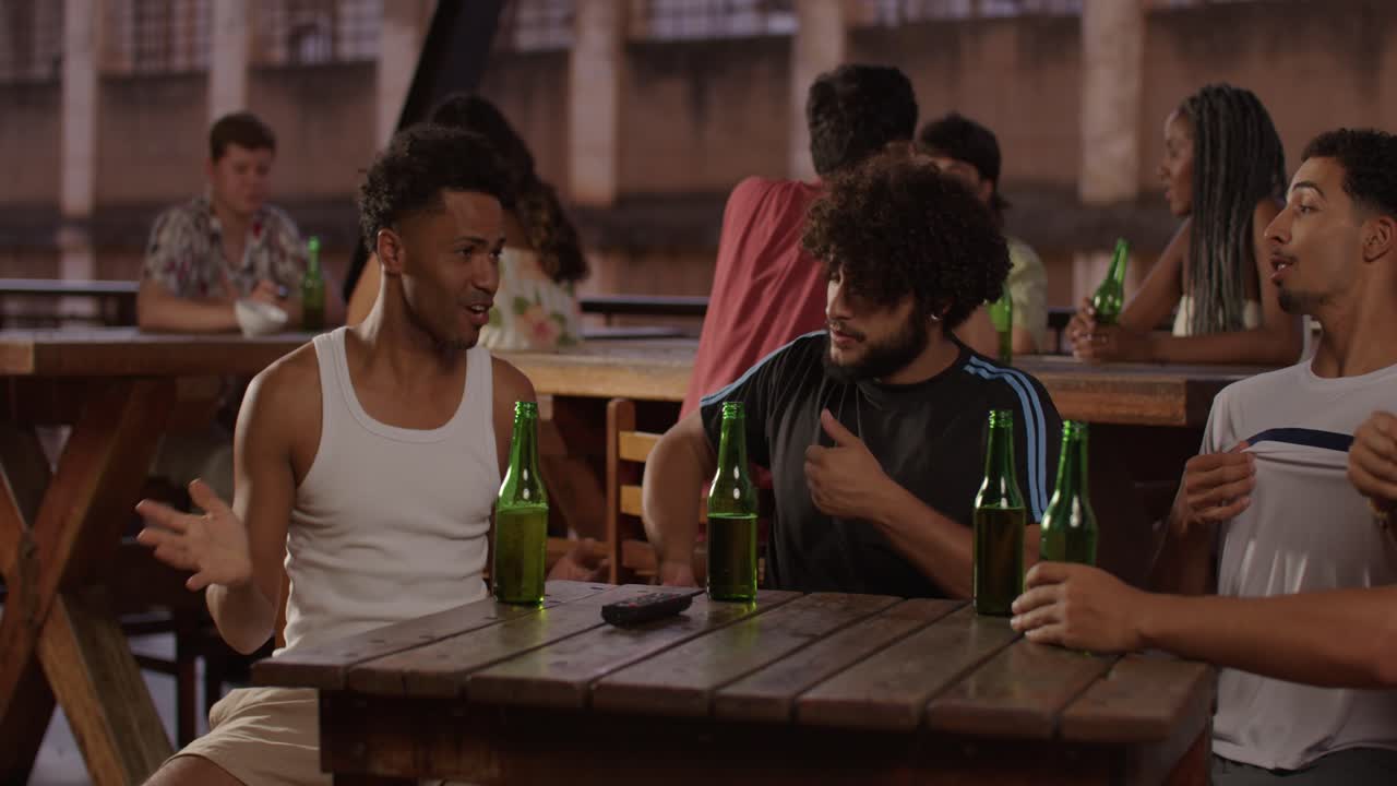 Friends socialising and drinking beer at a casual rooftop bar, with one man putting on a red shirt