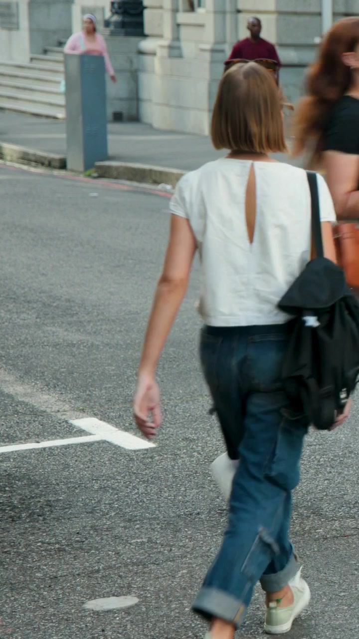 Vertical video: Opening shot showing woman in pink top striding across roadway, carrying backpack