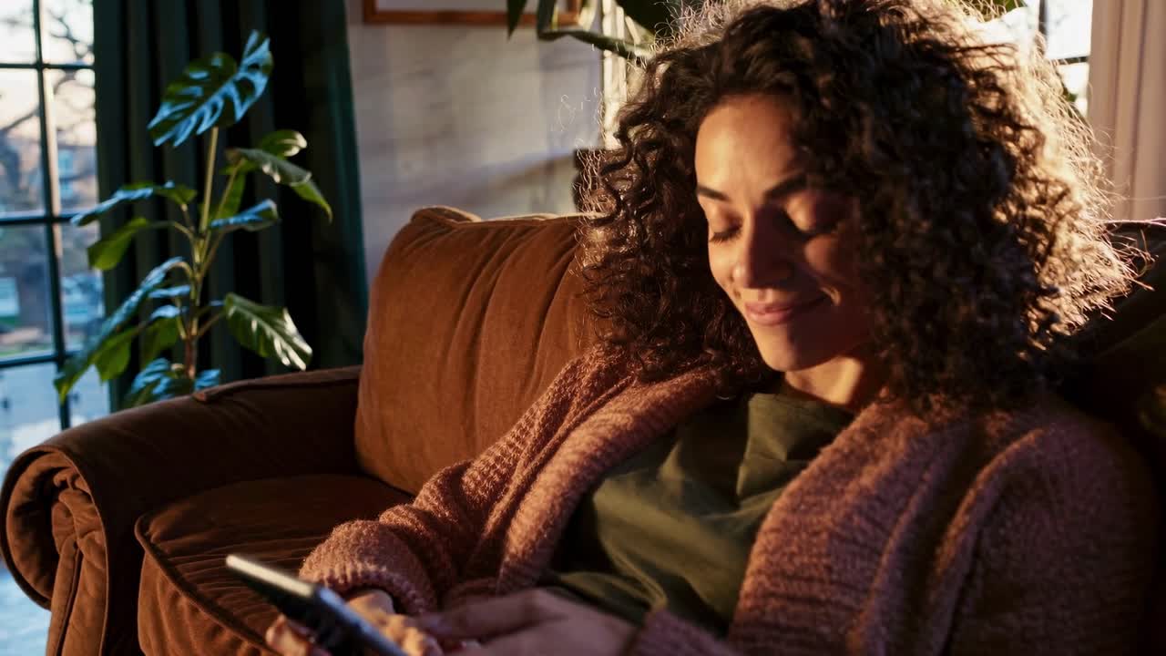 Una donna con i capelli ricci si rilassa su un divano, usando il suo telefono. Il video cattura un'atmosfera calda e accogliente.