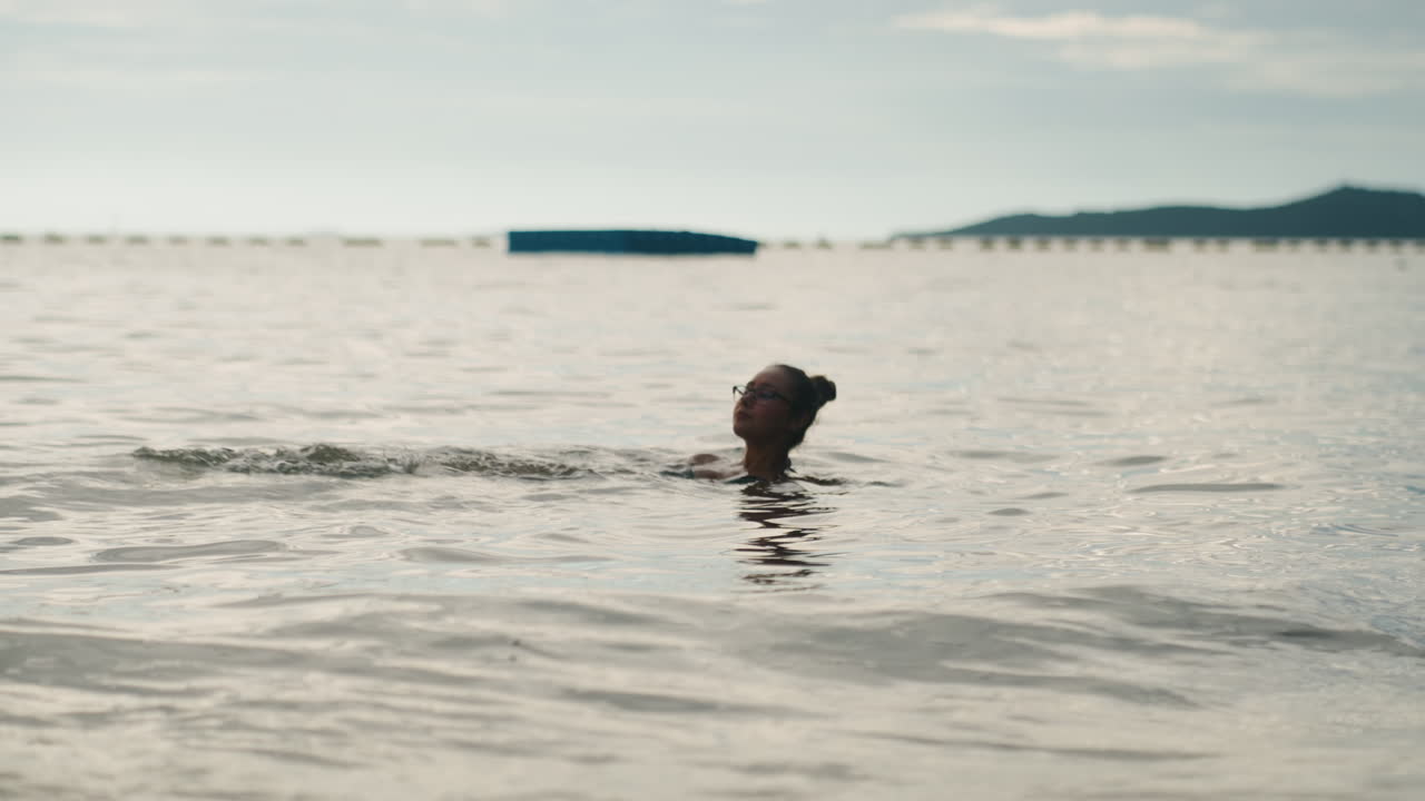 Solitary swimmer in calm sea, distant horizon closeup head above gentle ripples, buoy line and island silhouette on skyline, overcast dawn light, reflective mood, morning lap, therapeutic float