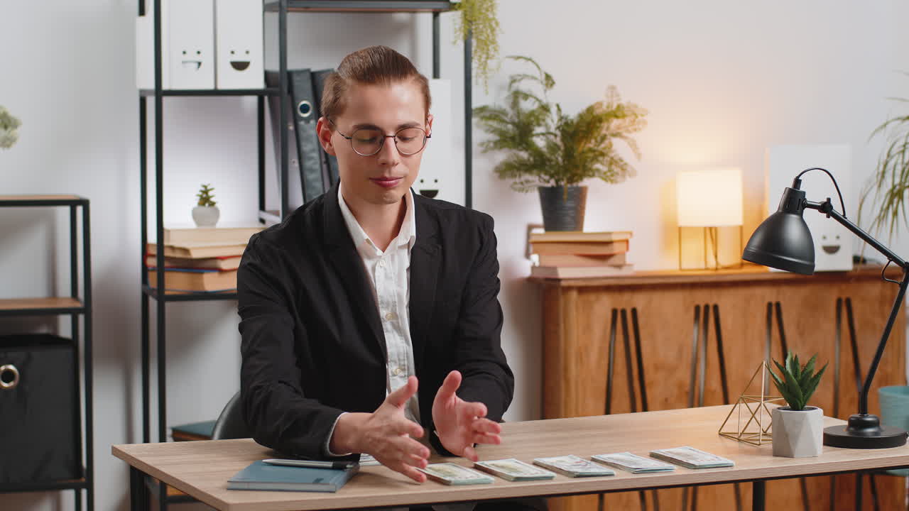 Successful young businessman guy in formal suit sitting counting money at table in home office