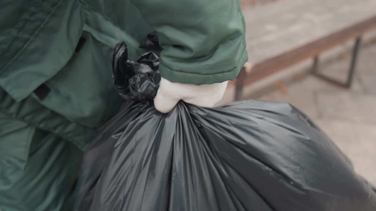 Worker lifting black trash bag on sidewalk with gloved hands and green uniform, hauling heavy sealed sack toward curb during routine city collection, visible boots and jacket suggesting cold weather