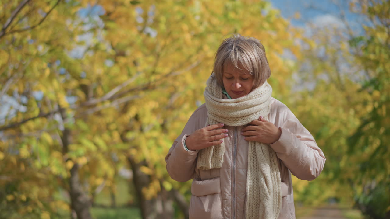 Mature woman in warm pink jacket and thick scarf walks confidently with hands tucked in pockets, adjusting muffler beneath golden autumn trees, surrounded by bright foliage and natural sunlight
