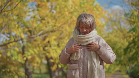 mature woman in warm pink jacket and thick scarf walks confidently with hands tucked in pockets, adjusting muffler beneath golden autumn trees, surrounded by bright foliage and natural sunlight