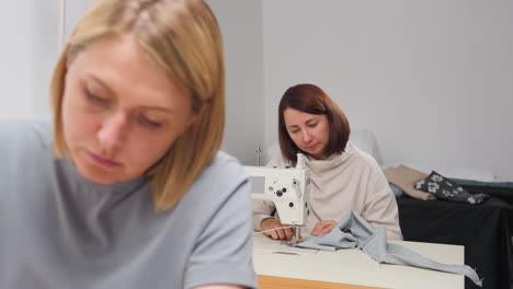 dress designers sit at sewing machines in bright workspace, concentrating on fabric stitching while textile pieces lie on table behind them, showing focus and creativity in professional setting
