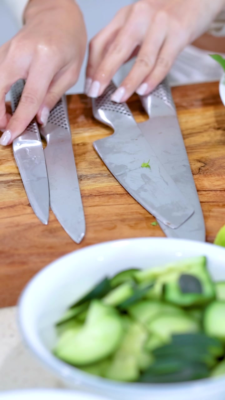 Eine Person ordnet Messer an und bereitet Gemüse für einen Salat auf einem Holzbrett in einer hellen Küche vor.