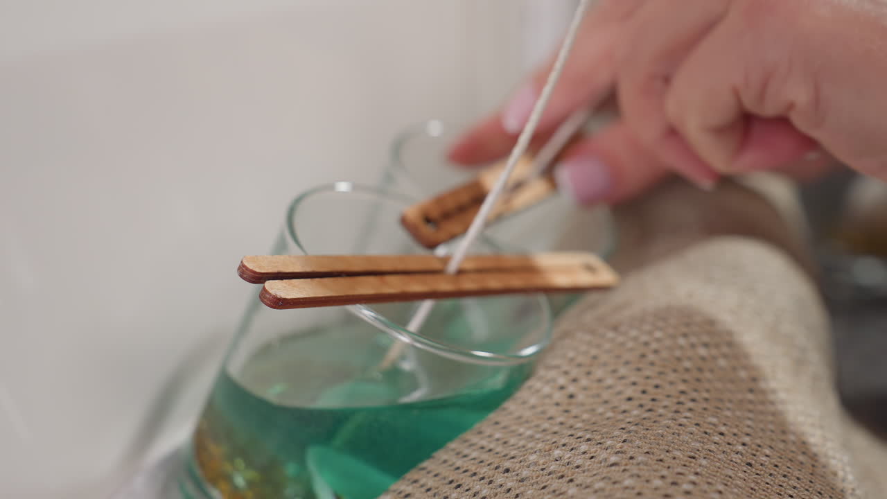 Close-up of glass cup with candle wick resting on sack bag, slight blur shows hands adjusting wick in second cup during candle-making process