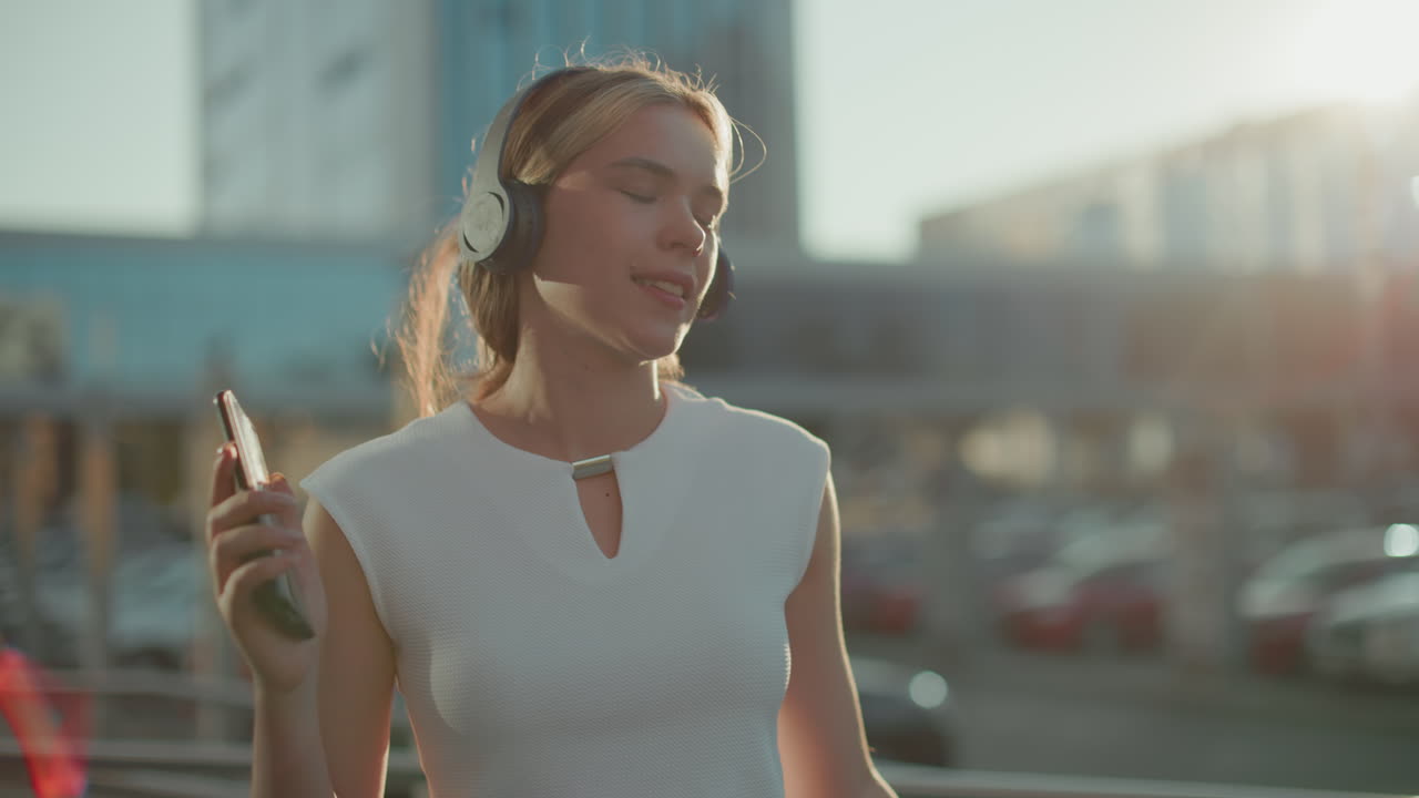 Golden sunlight ambiance surrounds young lady enjoying music with headphones and phone in hand while dancing gracefully in urban setting with slight blur background of cars and buildings