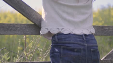 Carefree-woman-leans-on-countryside-gate-looking-at-field-of-rapeseed