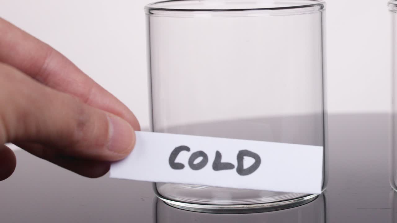 Hand places handwritten 'COLD' label on clear glass beaker, neutral lighting, stationary camera