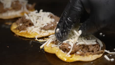 queso triturado añadido a los tacos de birria cocinar en una parrilla plana, manos negras con guantes salpicadas de quesadilla rallado a los taco chisporroteantes, deslizador en cámara lenta cerca de 4k