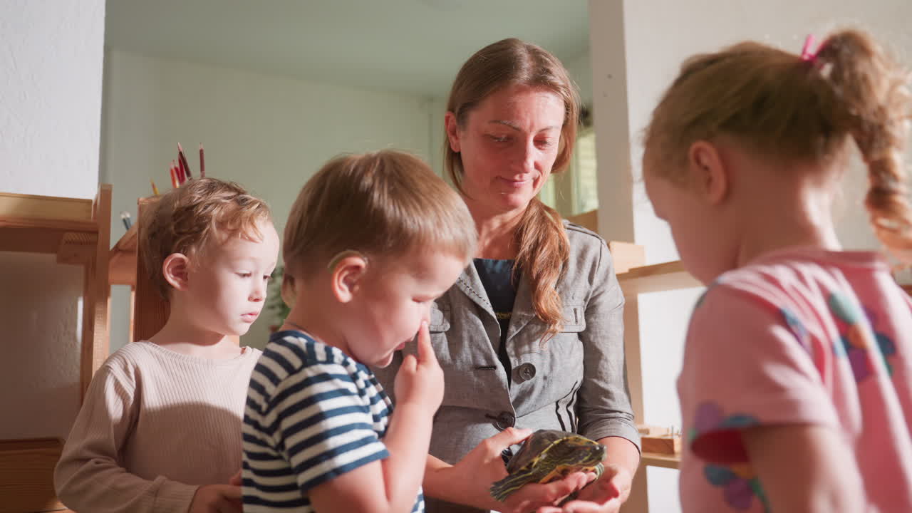 Ein Trainer stellt Kindern eine Schildkröte vor, während sie sich neugierig versammeln. Ein Kind mit Hörgerät und gestreiftem Hemd beobachtet aufmerksam, während andere die Interaktion in einer Unterrichtsumgebung konzentriert beobachten.