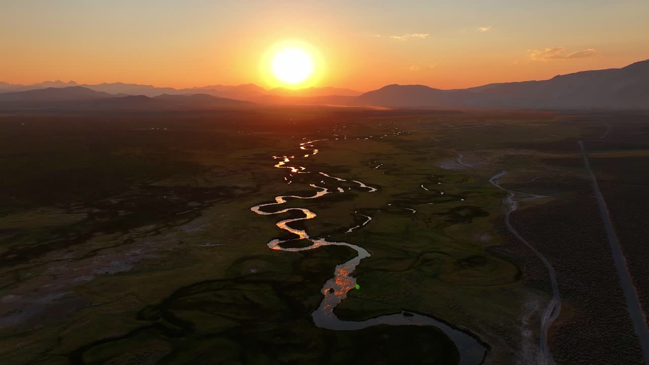 Drone tracks and orbits the setting sun over a meandering Owens River near Mammoth Lakes California. Golden hour light reflects across S curve channels and open valley with distant Sierra peaks