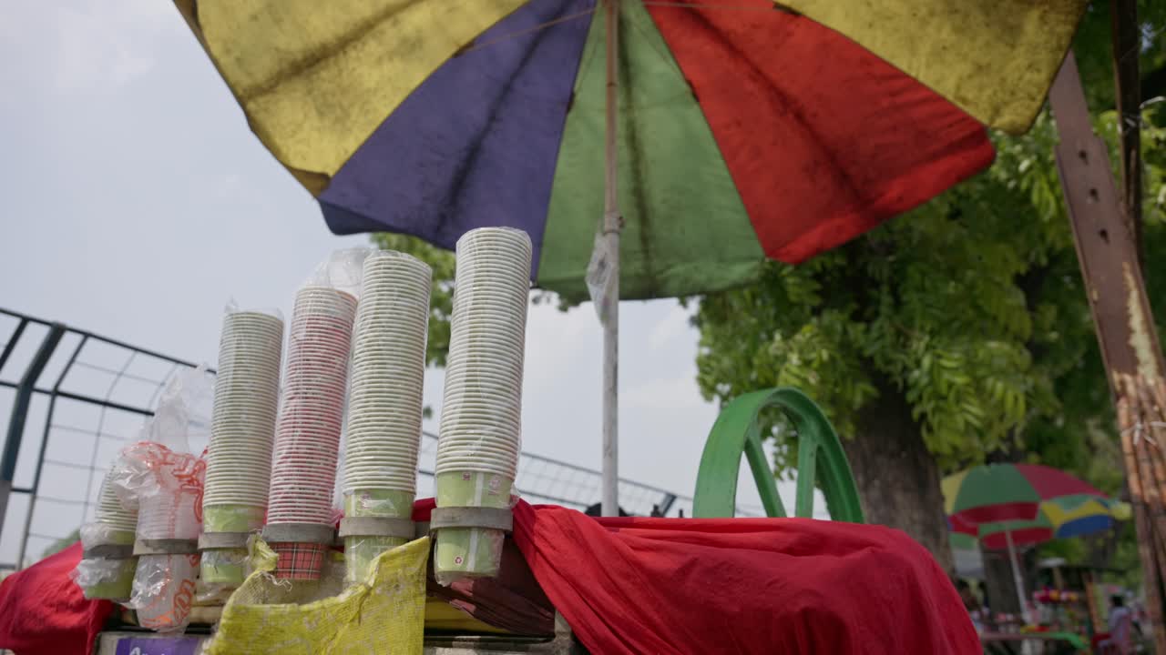 Nærbillede af stablede engangskrus ved en juicebod langs vejen under en farverig paraply. Vinden blæser, gadebillede og hverdagsmadscene på et indisk udendørs marked.