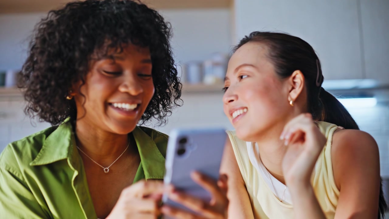 Female friends browsing phone home kitchen. Women laughing scrolling together.