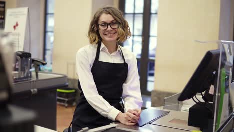 Woman-in-black-apron-as-a-cashier-at-the-cash-register-in-the-supermarket-or-discounter.-Smiling-short-haired-curly-woman